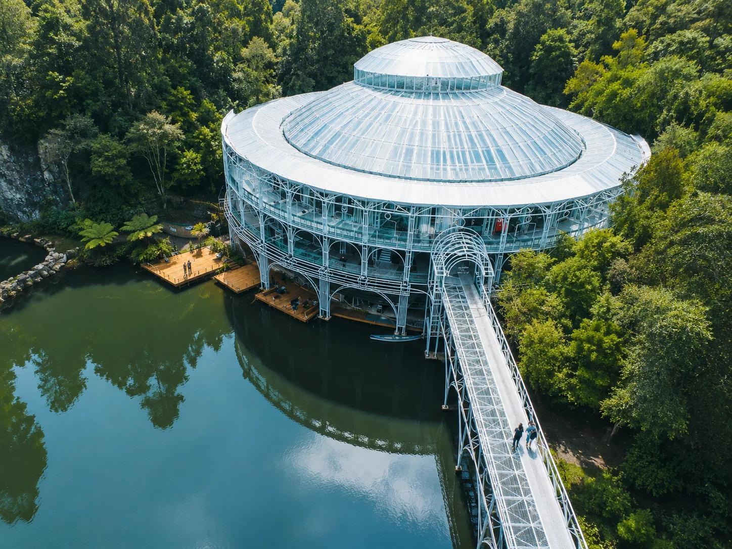 Vista aérea da Ópera de Arame, estrutura de vidro sobre o lago com a passarela, em Curitiba