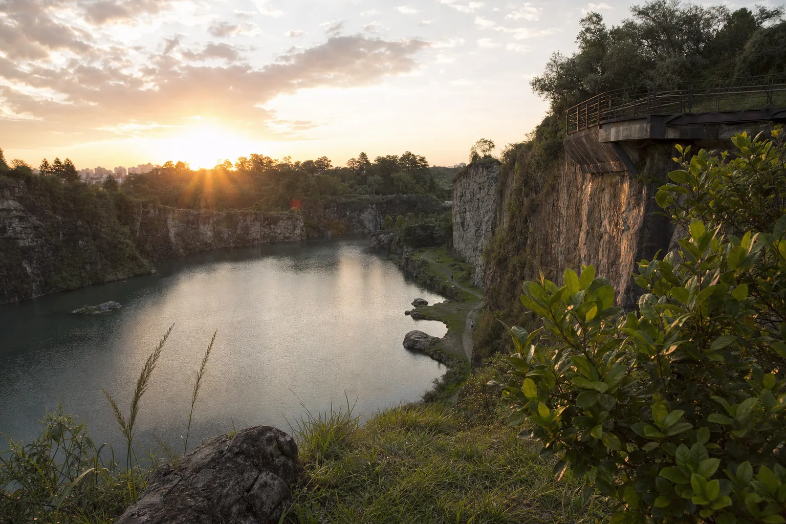 Parque em antigas pedreiras com paredões de pedra, lago e mirante ao pôr do sol, evocando o Parque Tanguá em Curitiba